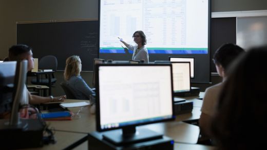 Students in a classroom with a teacher pointing to a blackboard