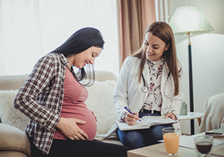 A pregnant woman sits on a couch while a midwife writes notes on a clipboard.